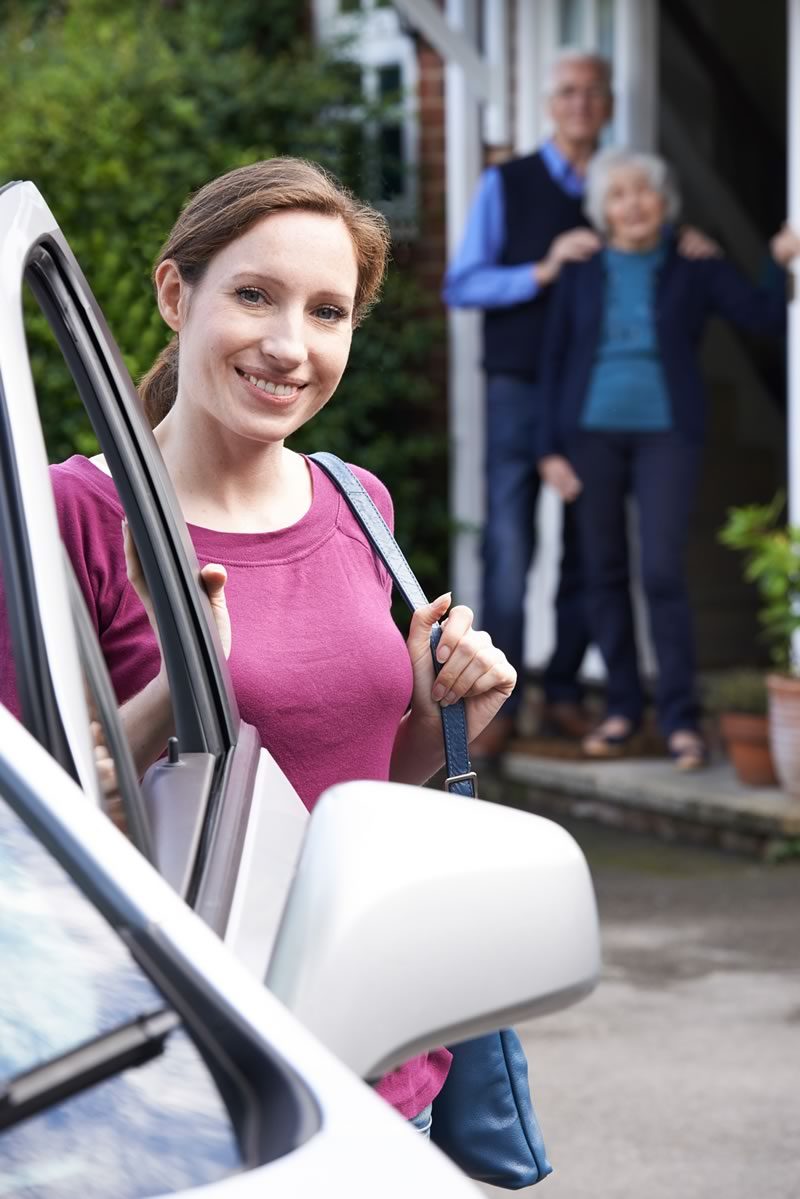 Young woman smiling, standing behind a car door while smiling parents are at their doorstep. 