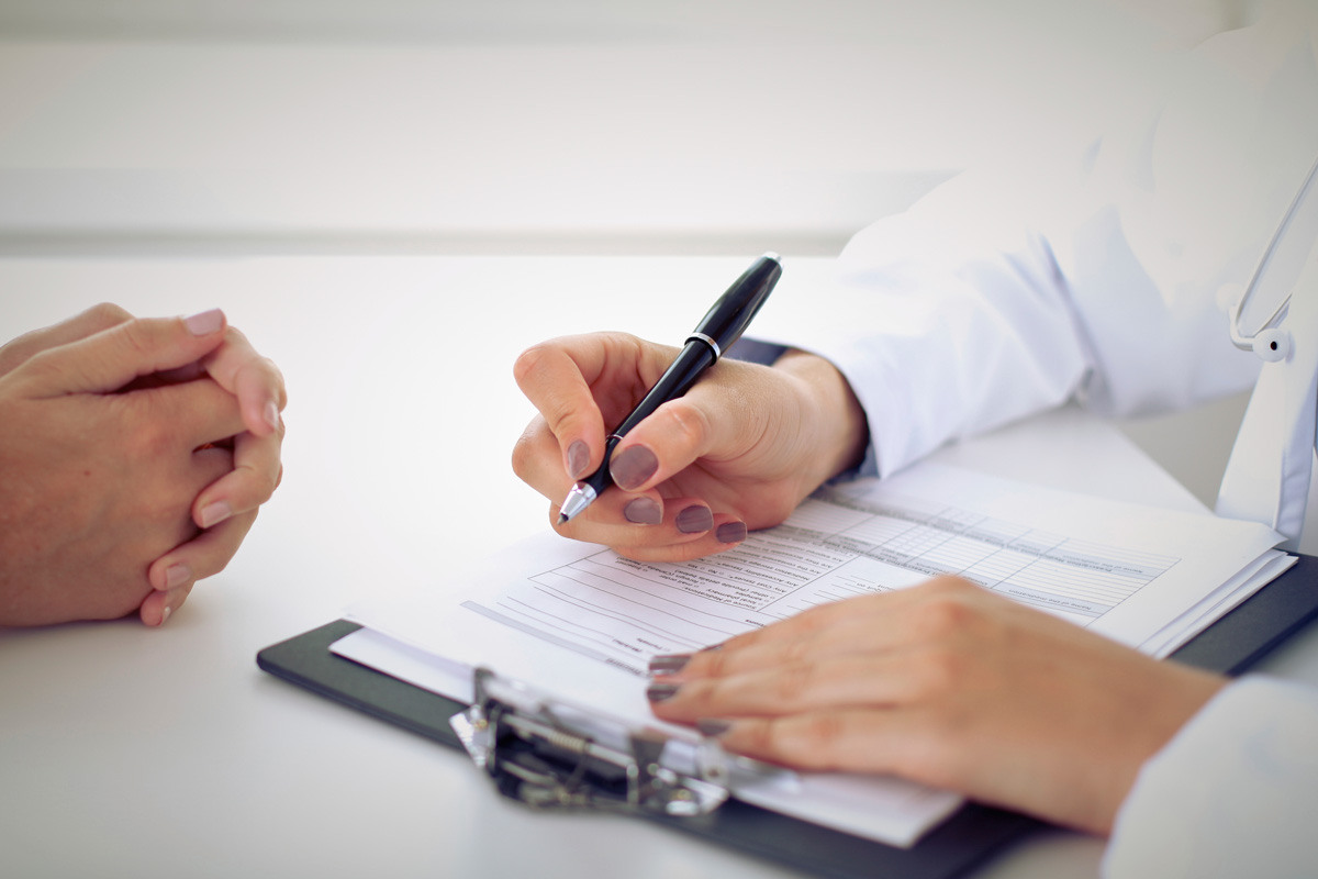Doctor's hands holding a pen and a clipboard with patient's hands clasped together across from the doctor. 