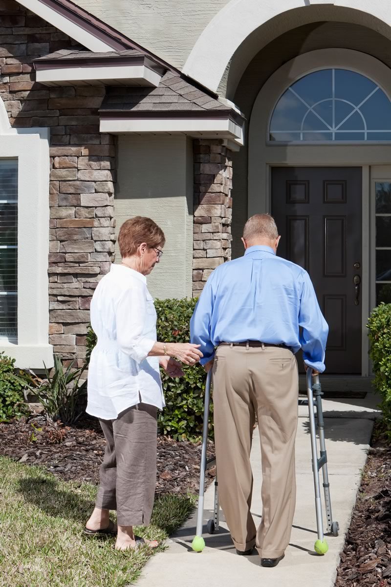 Senior couple walking into their home, one of them is using a walker, the other is supporting them. 